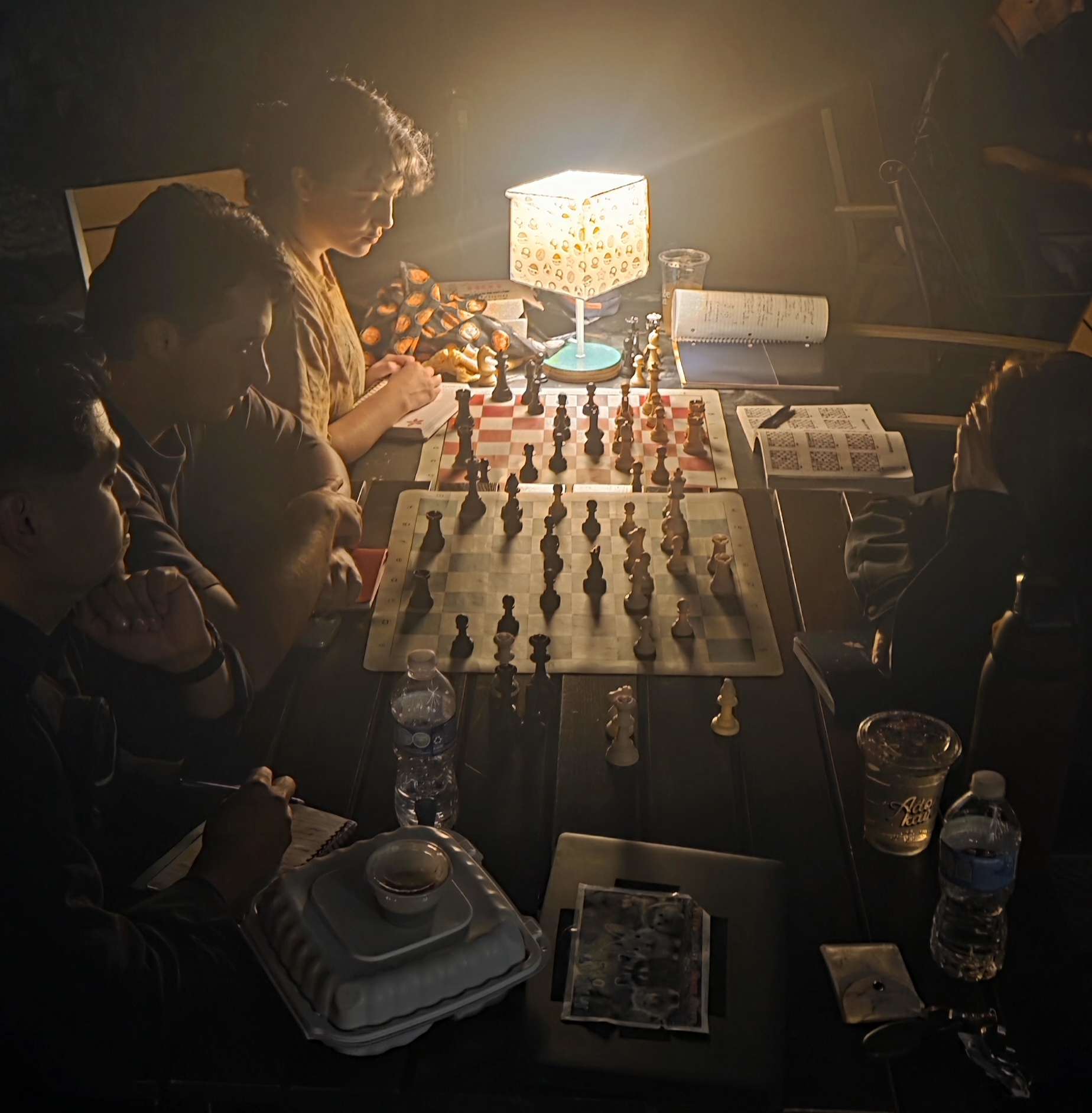 Jugadores resolviendo ejercicios tácticos a la luz de una lámpara en Adobe Kava, dos tableros y un libro de ejercicios abierto sobre la mesa