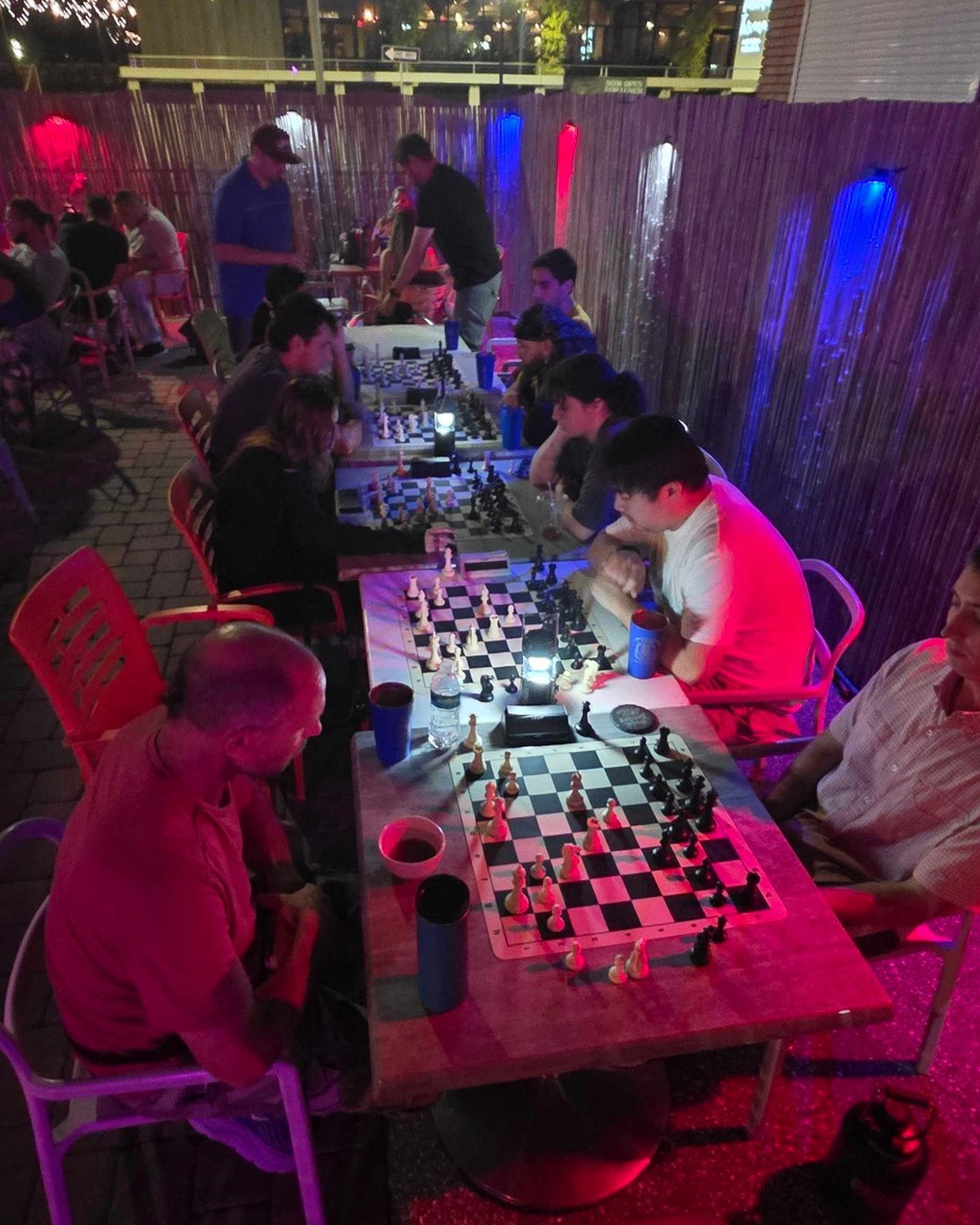 A long table of boards on the patio at Kava Social Club on a Tuesday study night, twelve players under the red and blue lights