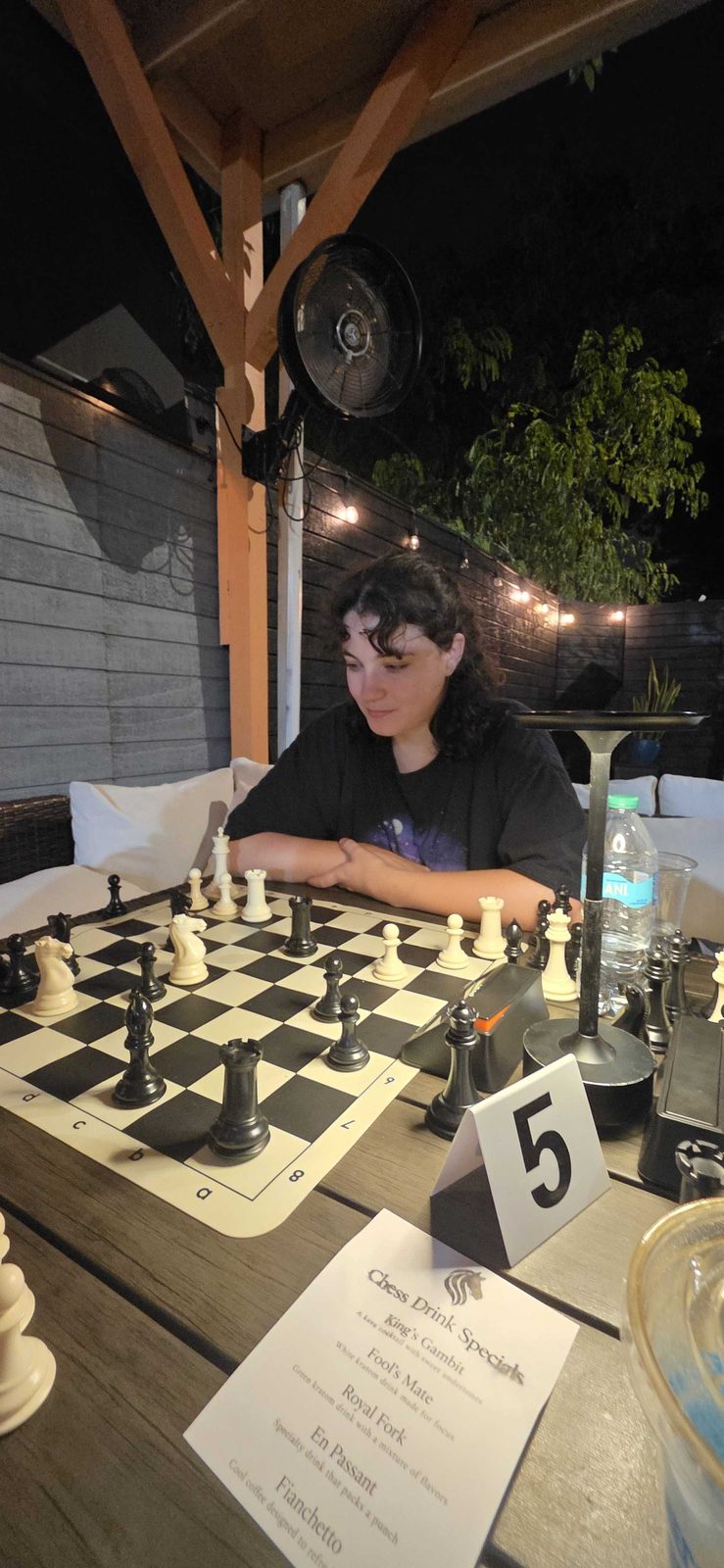 A player studying the board under the string lights, with the chess drink specials menu in front