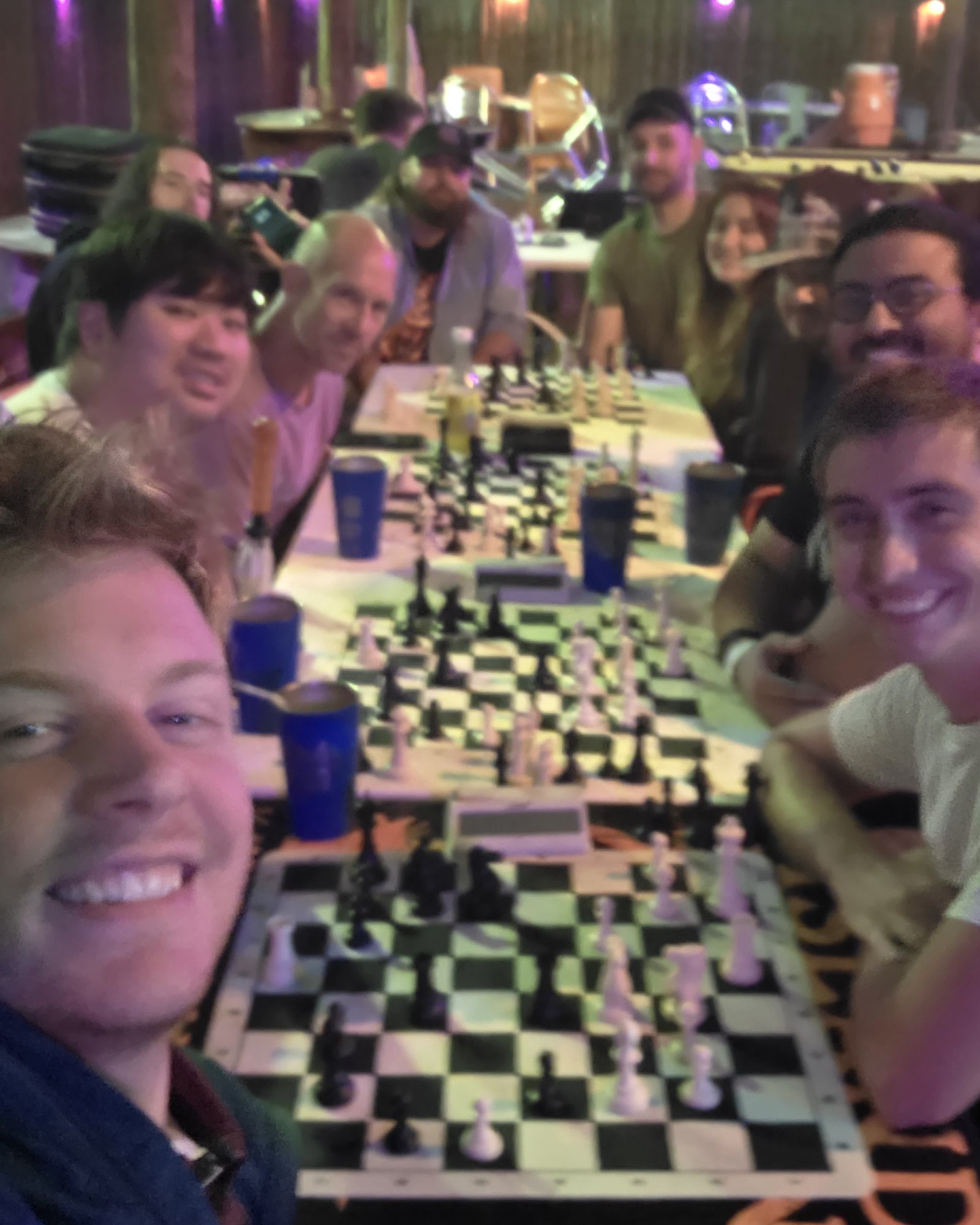 A smiling selfie down the long table under the tiki at Kava Social on a rainy study night, players and boards end to end
