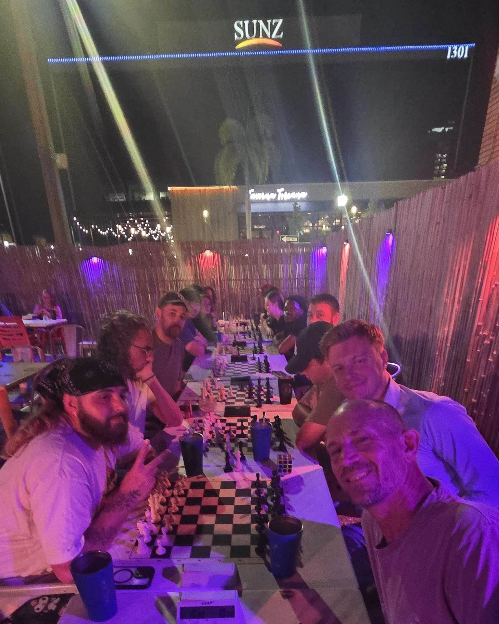 A smiling selfie down the long table on the patio under the SUNZ sign, players at their boards