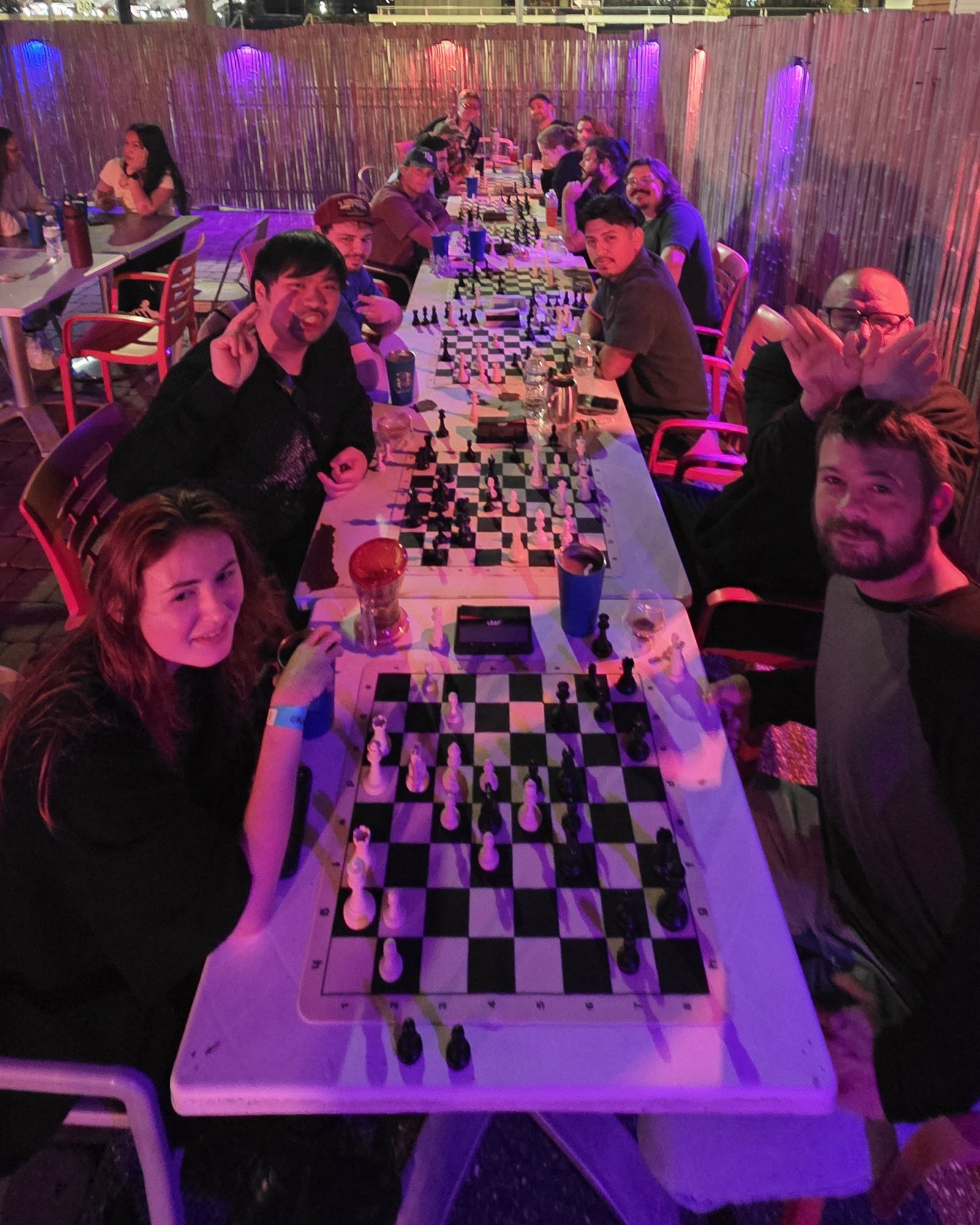 A full long table on the back patio at Kava Social, players turning to the camera from their boards under purple and red lights
